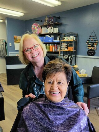 Jeanie Hertz standing behind client Janie Beasley in her chair with protective wrap on.