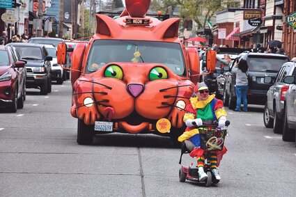 A van resembling an orange cat follows a clown driving a mobility scooter